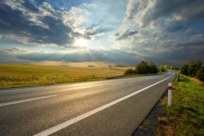 A rural highway is seen as the sun shines through the clouds above