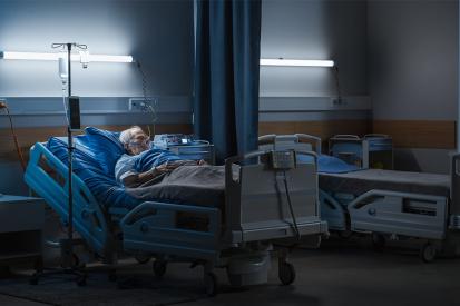 An elderly man lies asleep in bed in a dark hospital room