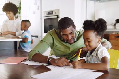 A father helps his daughter with homework while the rest of the family prepares food in the kitchen