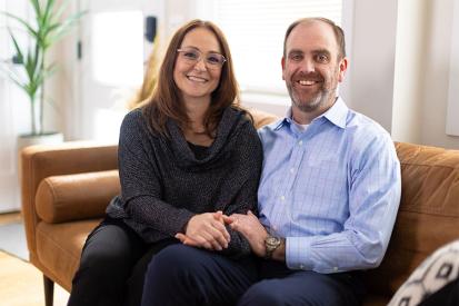 Joe and Serena Wailes sitting on a couch holding hands