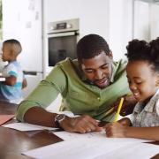 A father helps his daughter with homework while the rest of the family prepares food in the kitchen