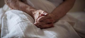 An elderly woman is seen in bed with her hands folded over her lap