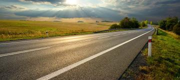 A rural highway is seen as the sun shines through the clouds above