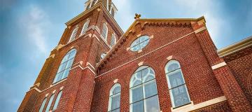 A brick church is seen on a bright sunny day with clouds behind the steeple