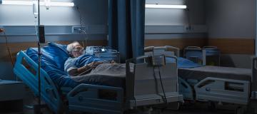 An elderly man lies asleep in bed in a dark hospital room