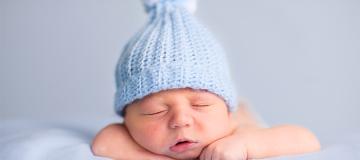 A baby wearing a blue beanie sleeps on a blanket