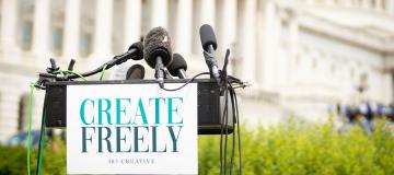 Microphones sit on a podium with a sign that reads 'Create Freely' in front of the U.S. Capitol