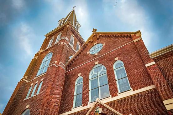 A brick church is seen on a bright sunny day with clouds behind the steeple