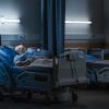 An elderly man lies asleep in bed in a dark hospital room